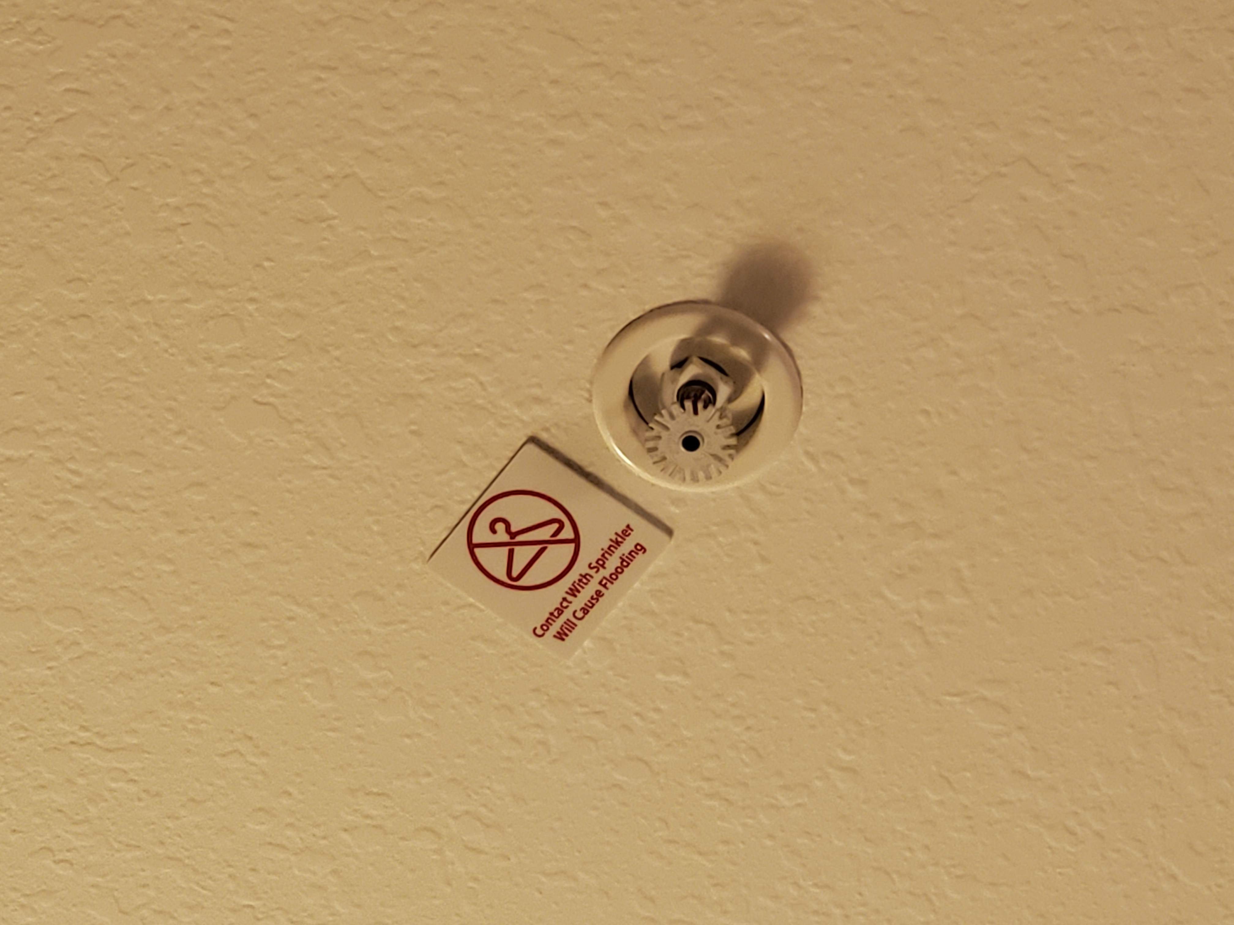 A hotel ceiling with a fire sprinkler and warning sticker showing a crossed-out coat hanger. The text reads: Contact With Sprinkler Will Cause Flooding.